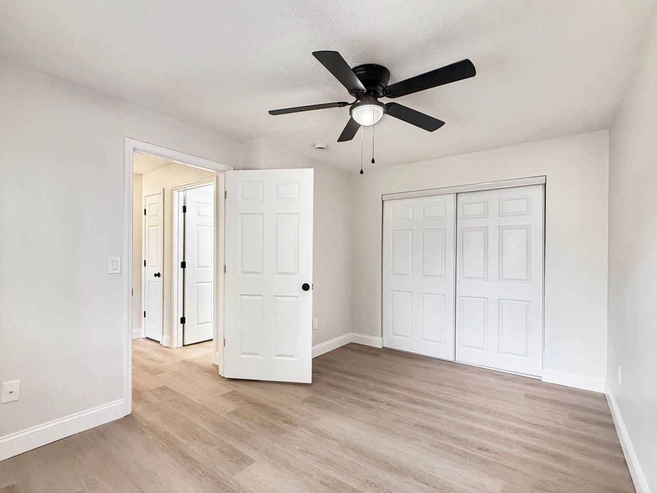 Empty bedroom with white walls, ceiling fan, open door, and light wood flooring