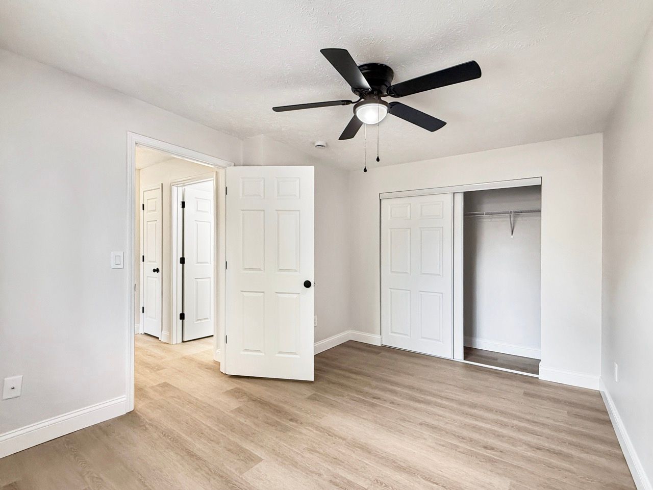 Empty white bedroom with light wood floor, ceiling fan, and open closet doors