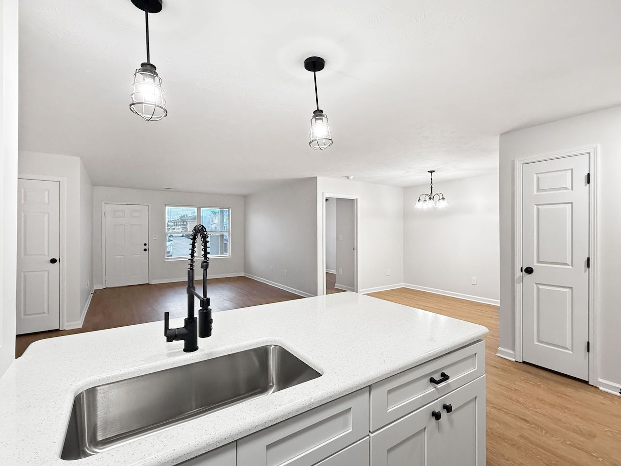 Bright kitchen with white cabinets, island sink, pendant lights, and hardwood floors opening to a dining area.