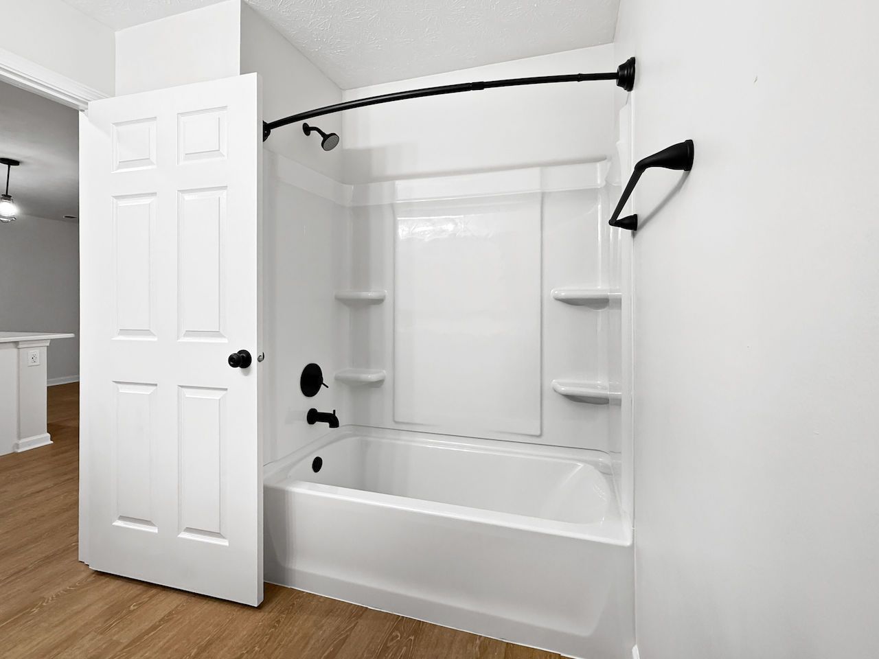 White bathroom with a bathtub, shower curtain rod, wall shelves, and an open door to the hall.