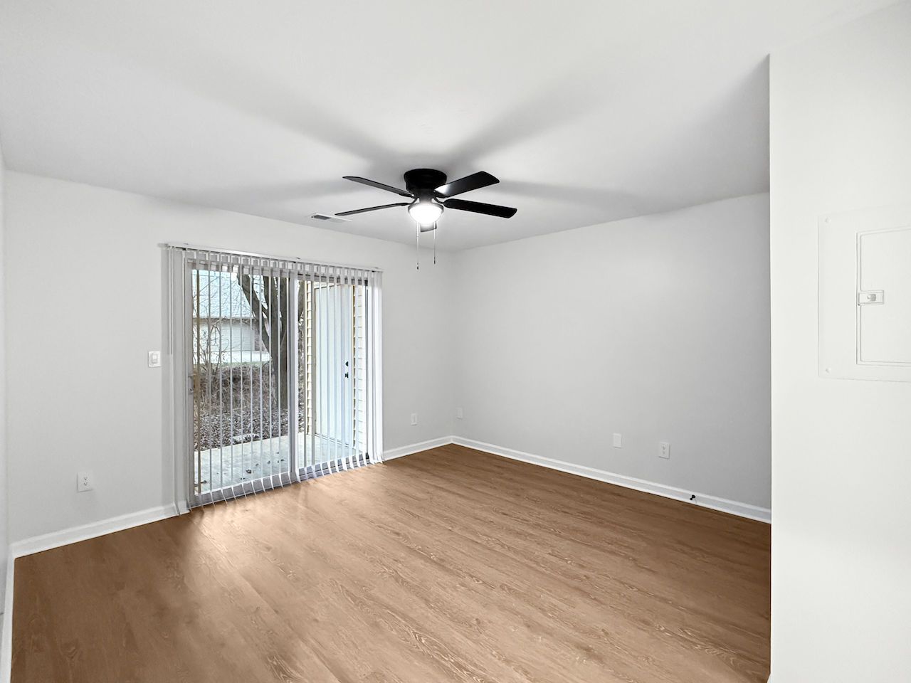 Empty living room with light gray walls, wood floors, ceiling fan, and sliding glass doors.