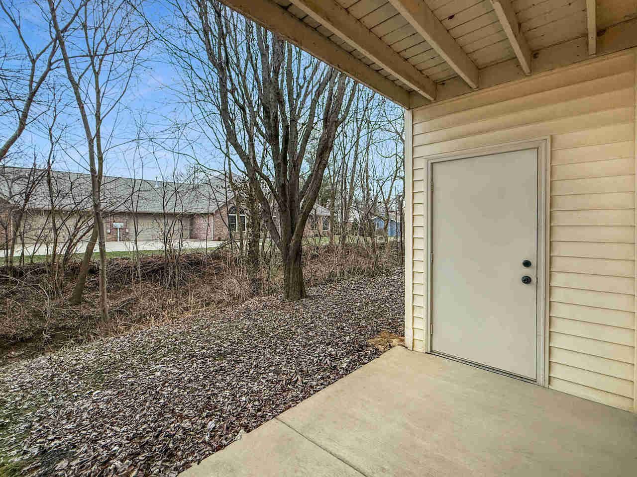 Covered patio with a beige door, concrete slab, and leaf-covered yard with trees beyond
