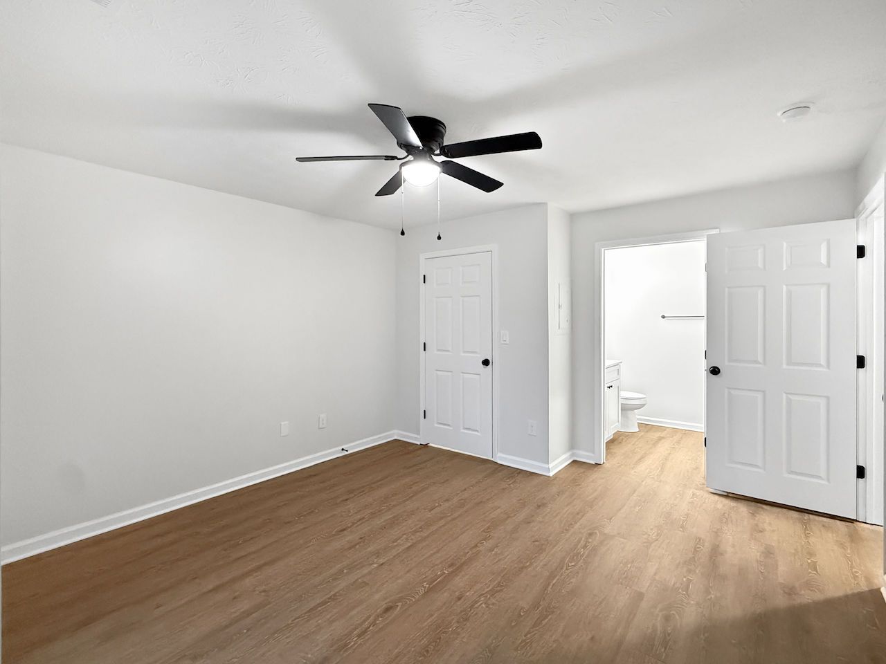 Empty white bedroom with wood floors, ceiling fan, closet doors, and open doorway to a hall