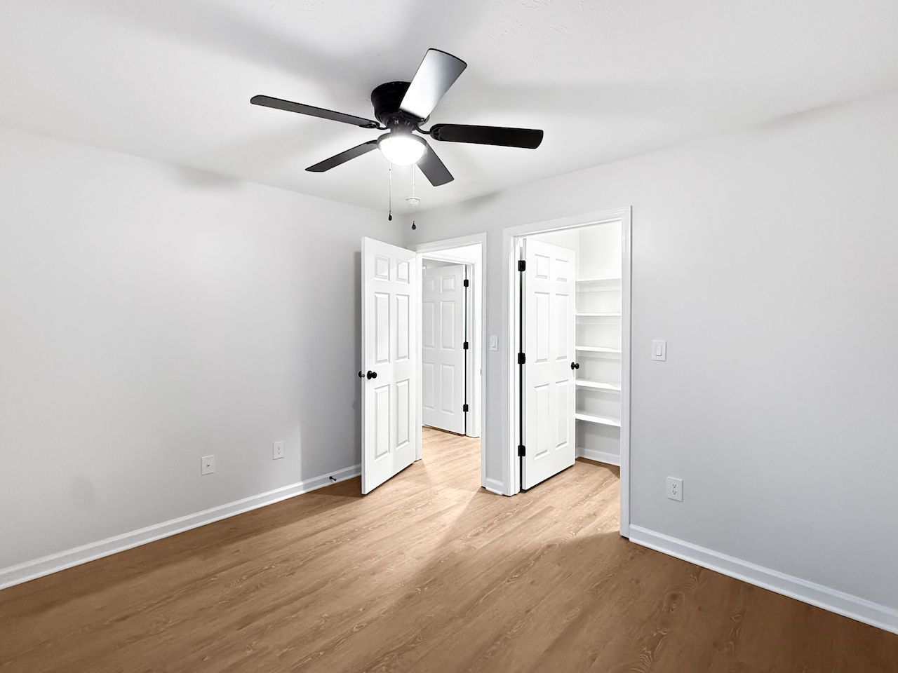 Empty white bedroom with ceiling fan, open double doors, and light wood flooring.