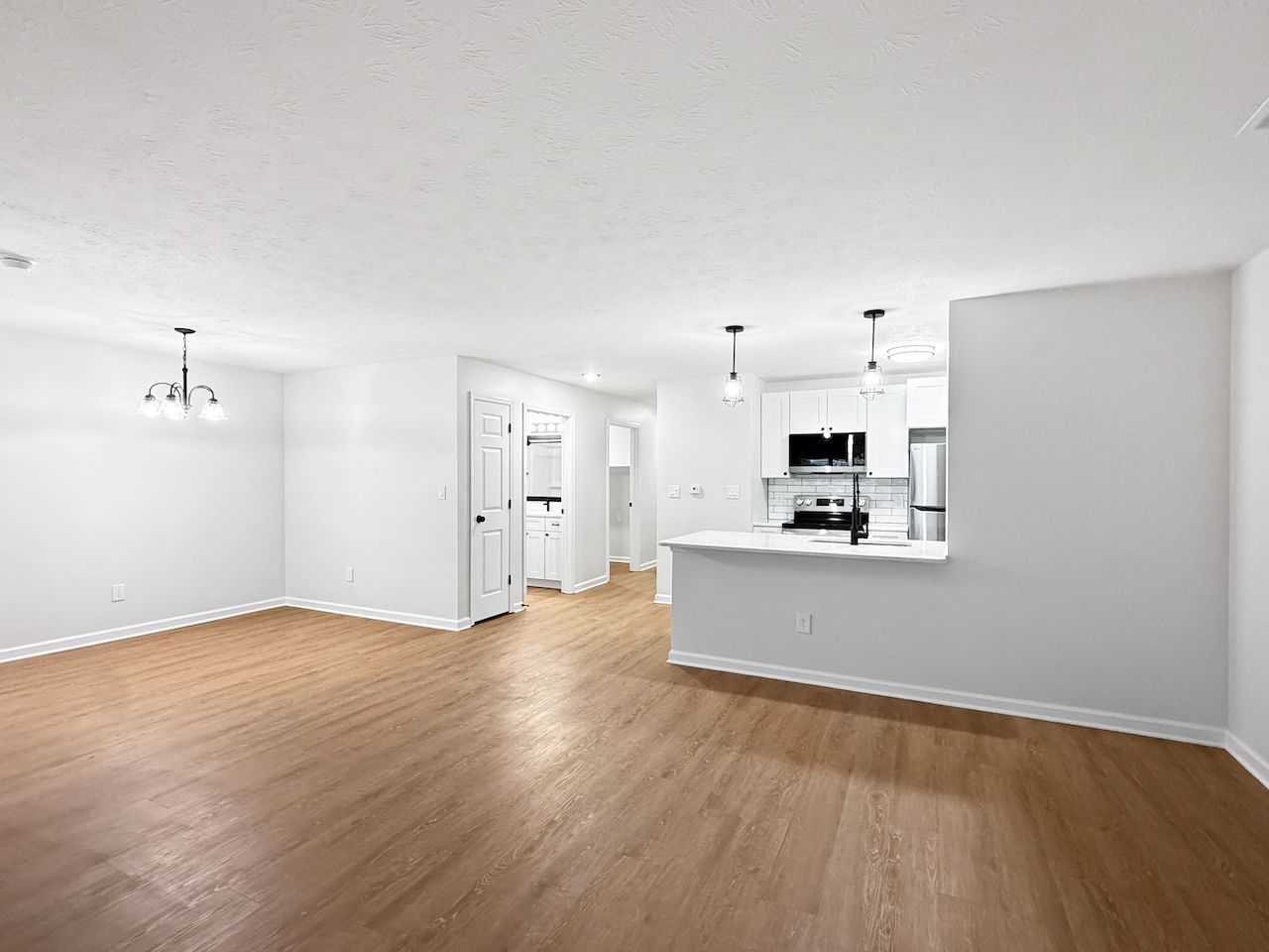 Empty open-plan living room with hardwood floors and white walls, opening to a bright kitchen with a breakfast bar
