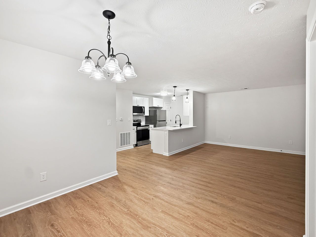 Empty open-plan living room with wood-look floor, white walls, and a chandelier near a kitchen pass-through.