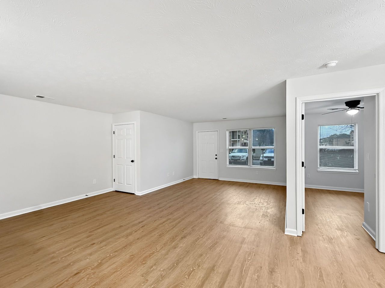 Empty bright living room with wood floors, white walls, and a doorway to a kitchen or hallway.