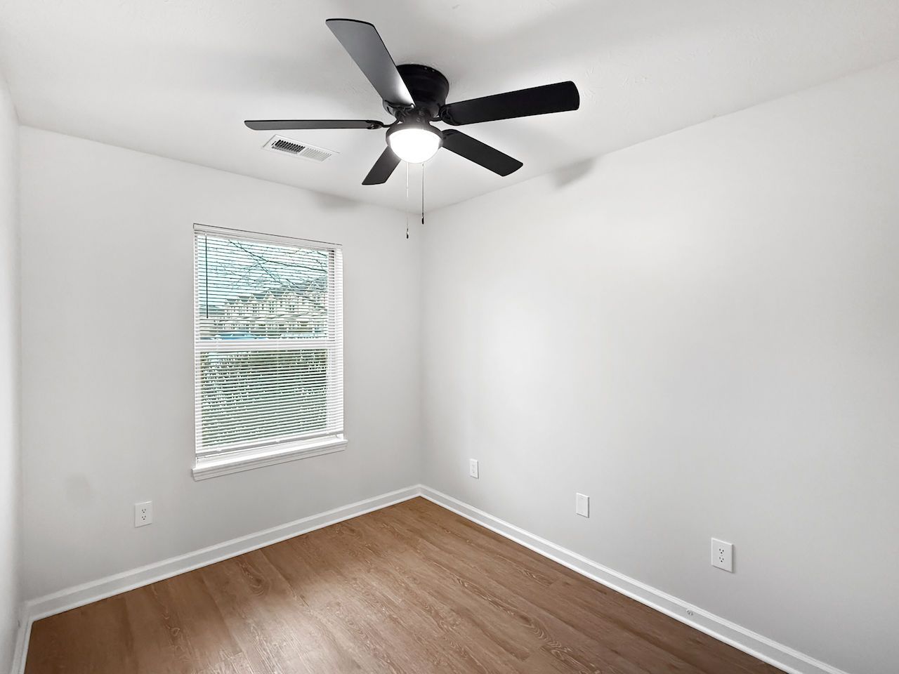 Empty white bedroom with wood floor, ceiling fan, and small window with blinds