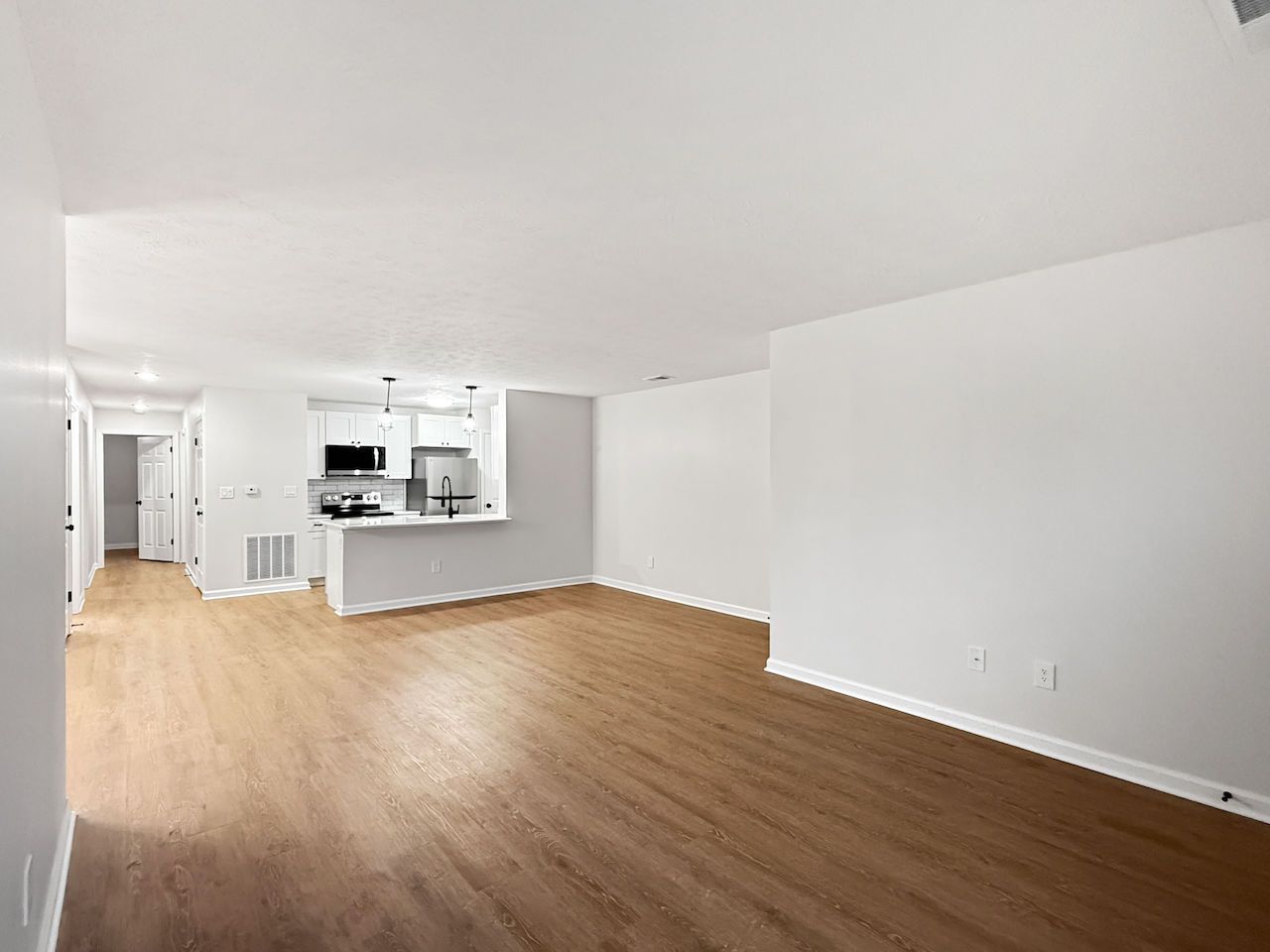 Empty modern living room with wood floors, white walls, and open kitchen pass-through.