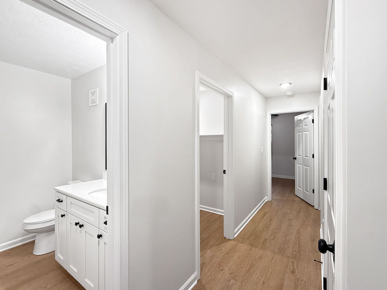 Bright hallway with white walls, wood floors, and a bathroom doorway on the left.