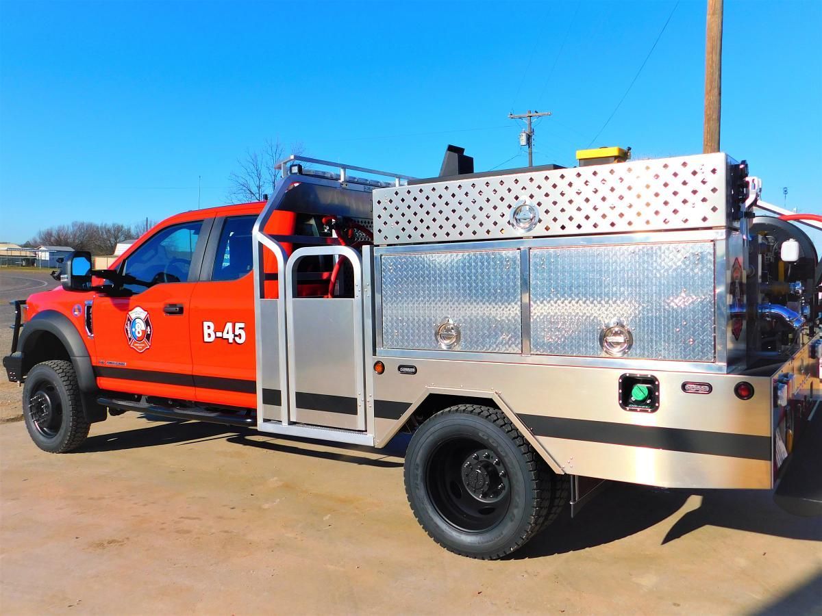 Orange fire rescue truck with silver equipment bed parked on a dirt lot under a blue sky
