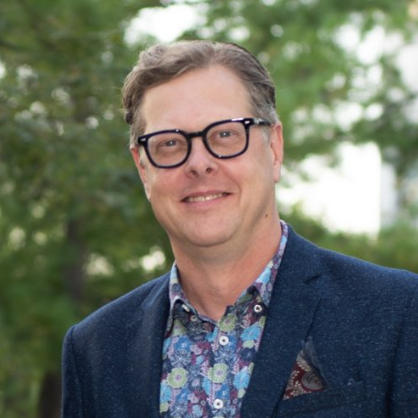 Man with glasses, leaning near a window, wearing a patterned shirt, smiling slightly.