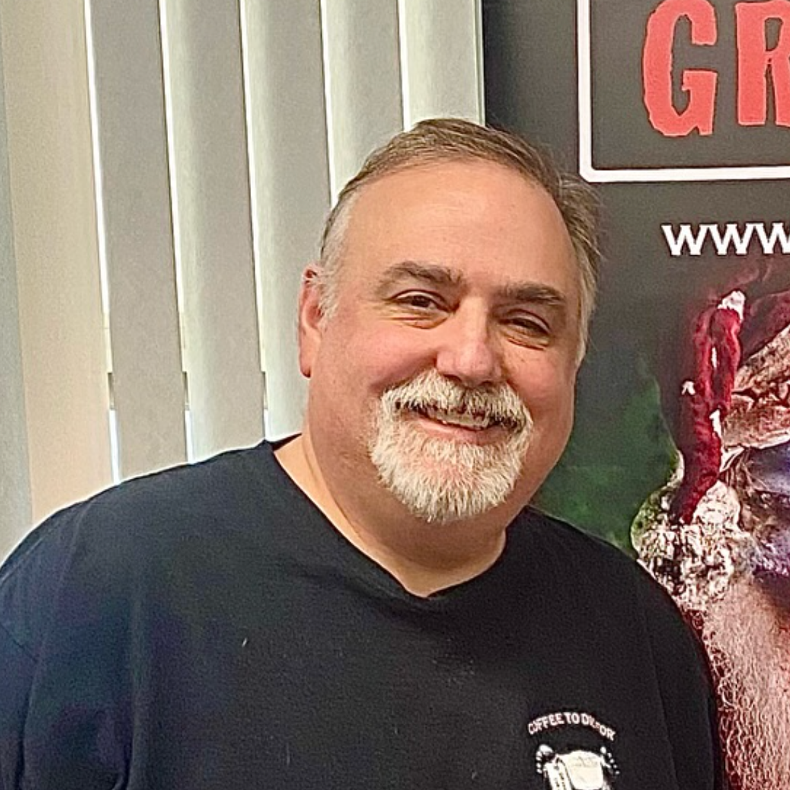 Man with a goatee smiles, wearing a black shirt, standing next to a poster.
