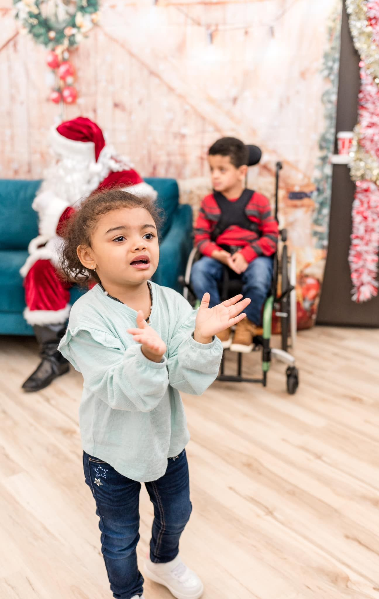 Girl clapping in front of Santa and a boy in a wheelchair. Festive background with wood and flowers.