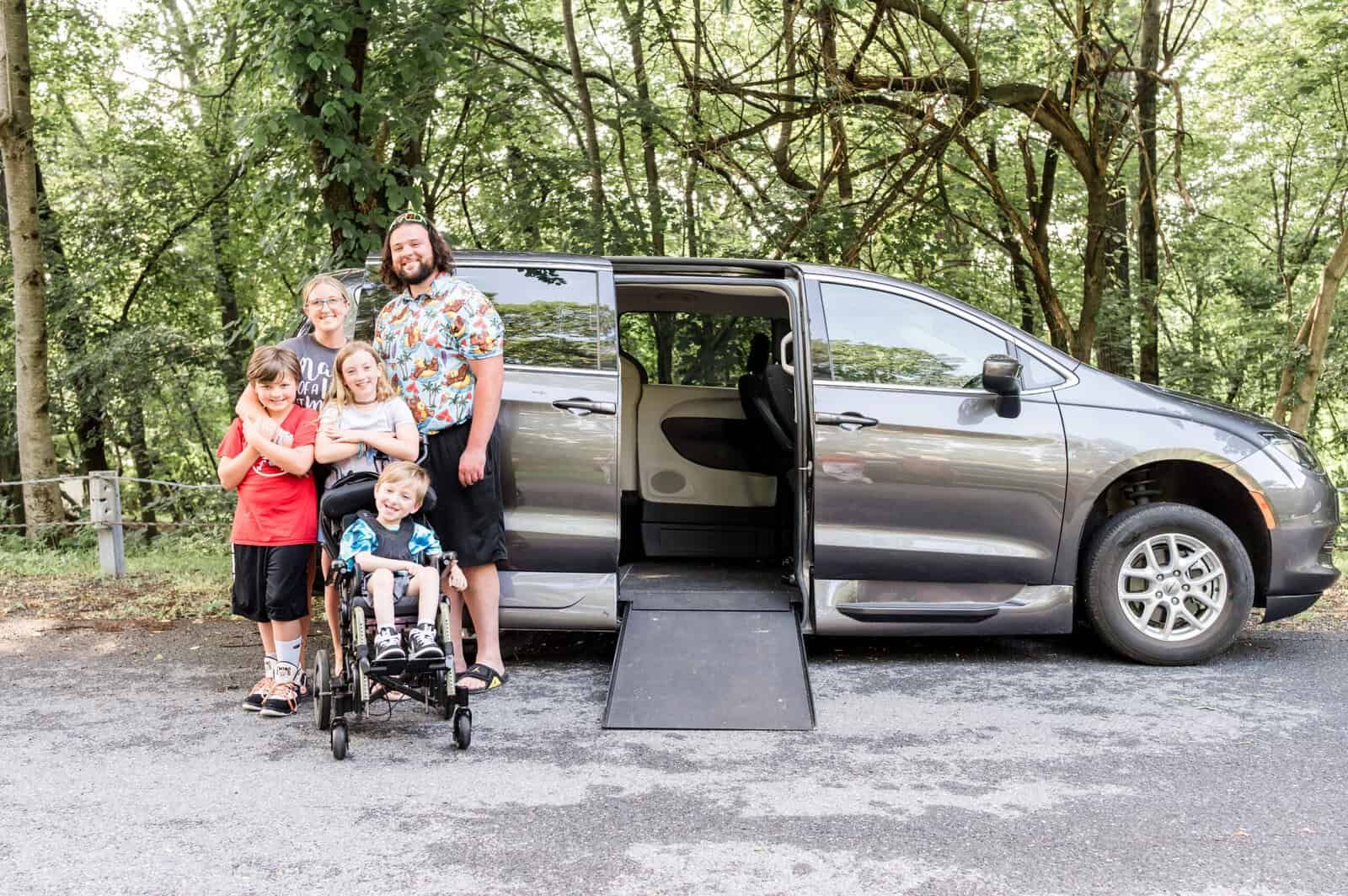Family stands beside a gray accessible van with a ramp. One child is in a wheelchair. Forest background.