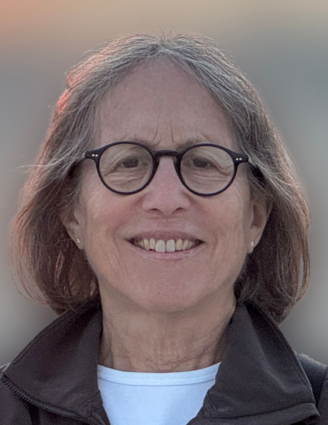 Smiling person with shoulder-length gray hair and round glasses, wearing a dark jacket outdoors.