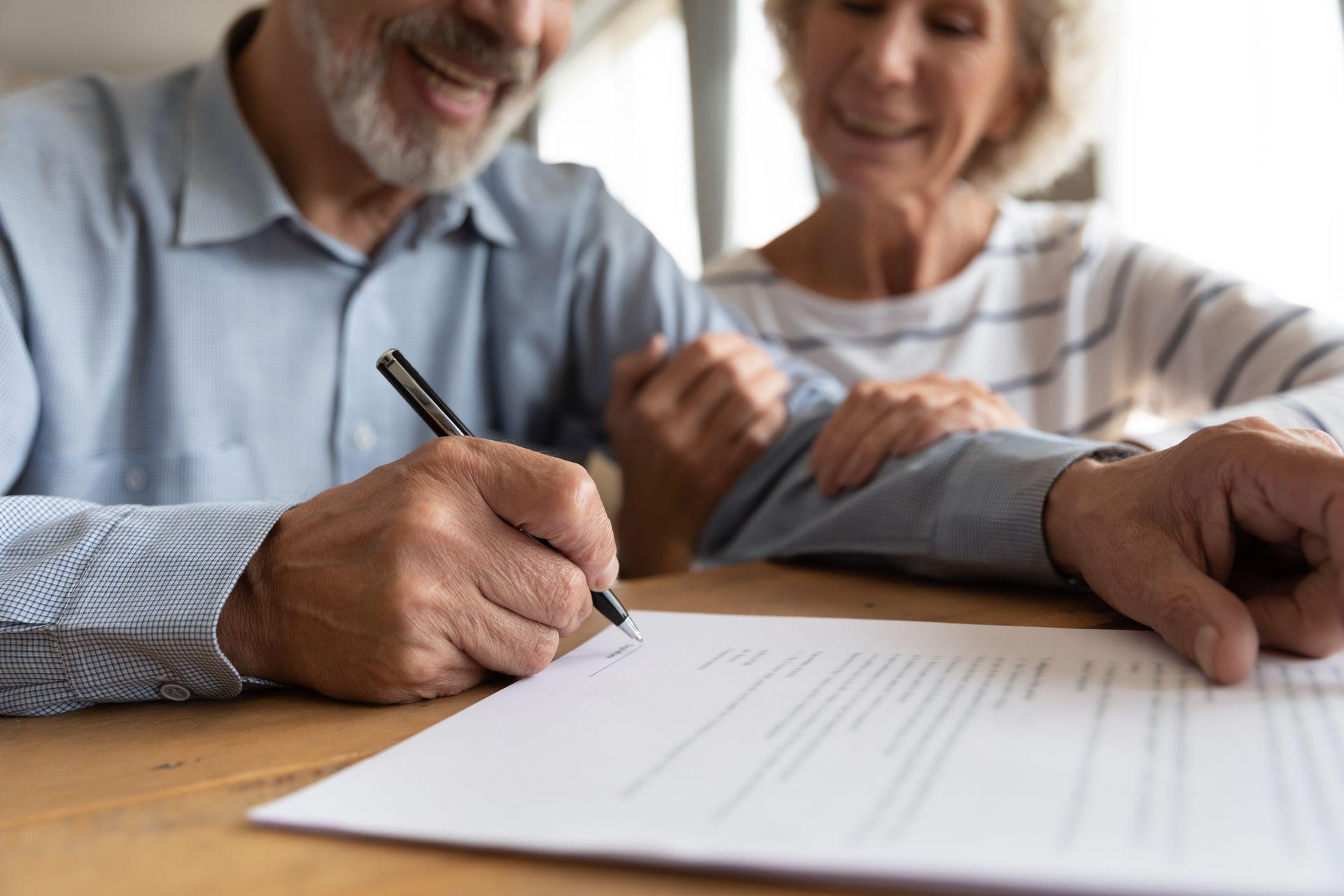 Senior man signing a document, woman with hand on his shoulder, smiling. Wooden table, indoors.