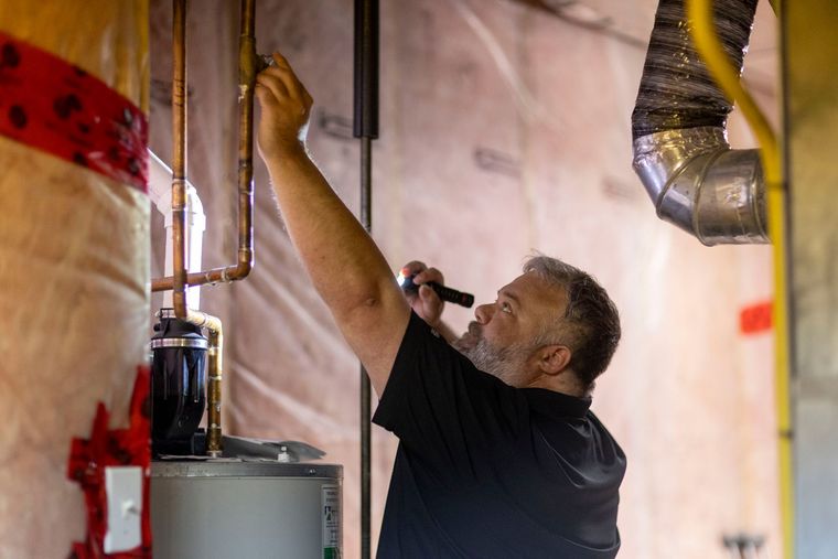 Man inspecting plumbing equipment in a utility room, reaching up toward exposed pipes and ducts.