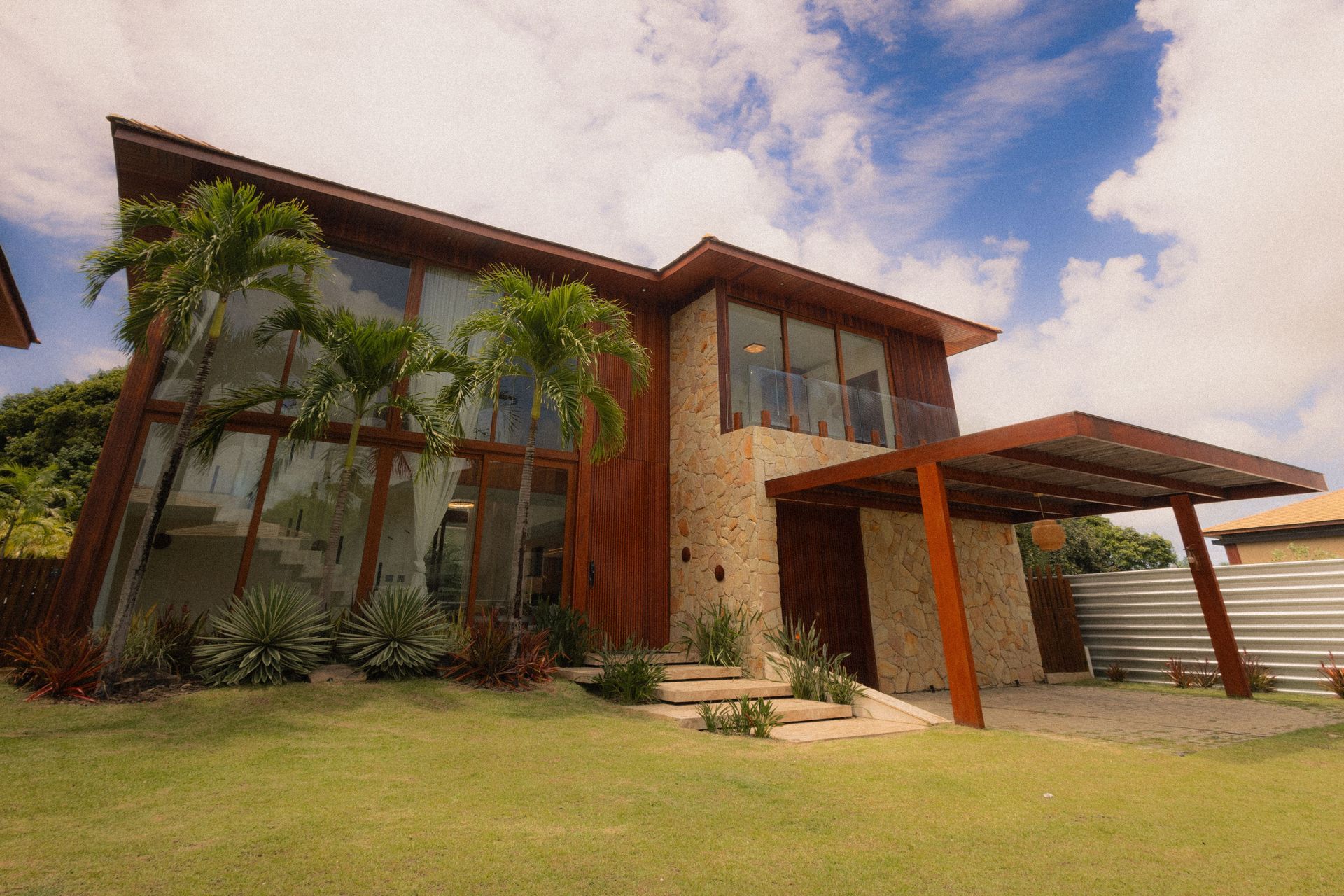 Modern two-story house with large windows, palm trees, and a covered driveway under a cloudy sky