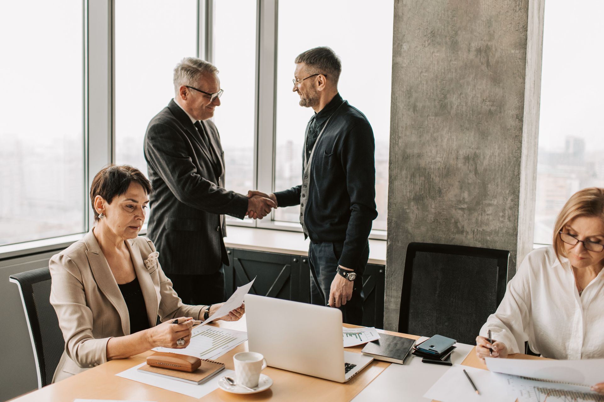 Colleagues in a bright office shaking hands while two others work at a table with laptops and papers
