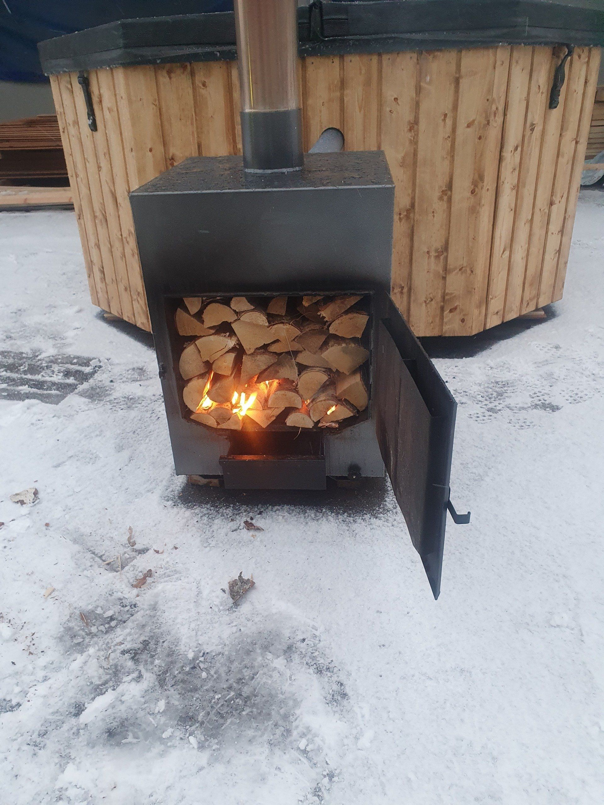 Brændeovn med åben dør og synlig ild, placeret på snedækket jord ved siden af ​​en trækonstruktion.