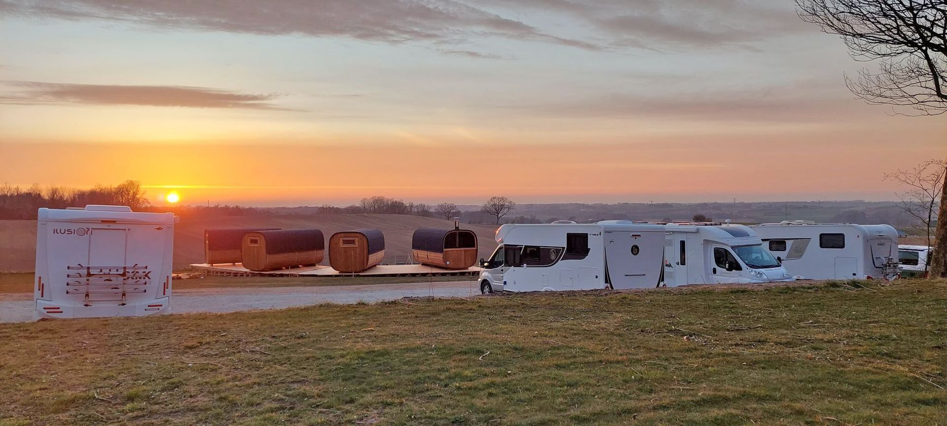Solnedgang over en græsklædt mark med rækker af hvide telte og parkerede varevogne i horisonten