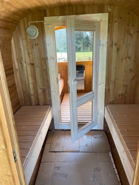 Sauna i træ med bænke, glasdør åben til et lille badeværelse og udsigt udover.