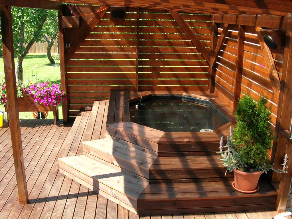 Træboblebad på en solbeskinnet terrasse under en pergola med potteplanter og trapper i nærheden