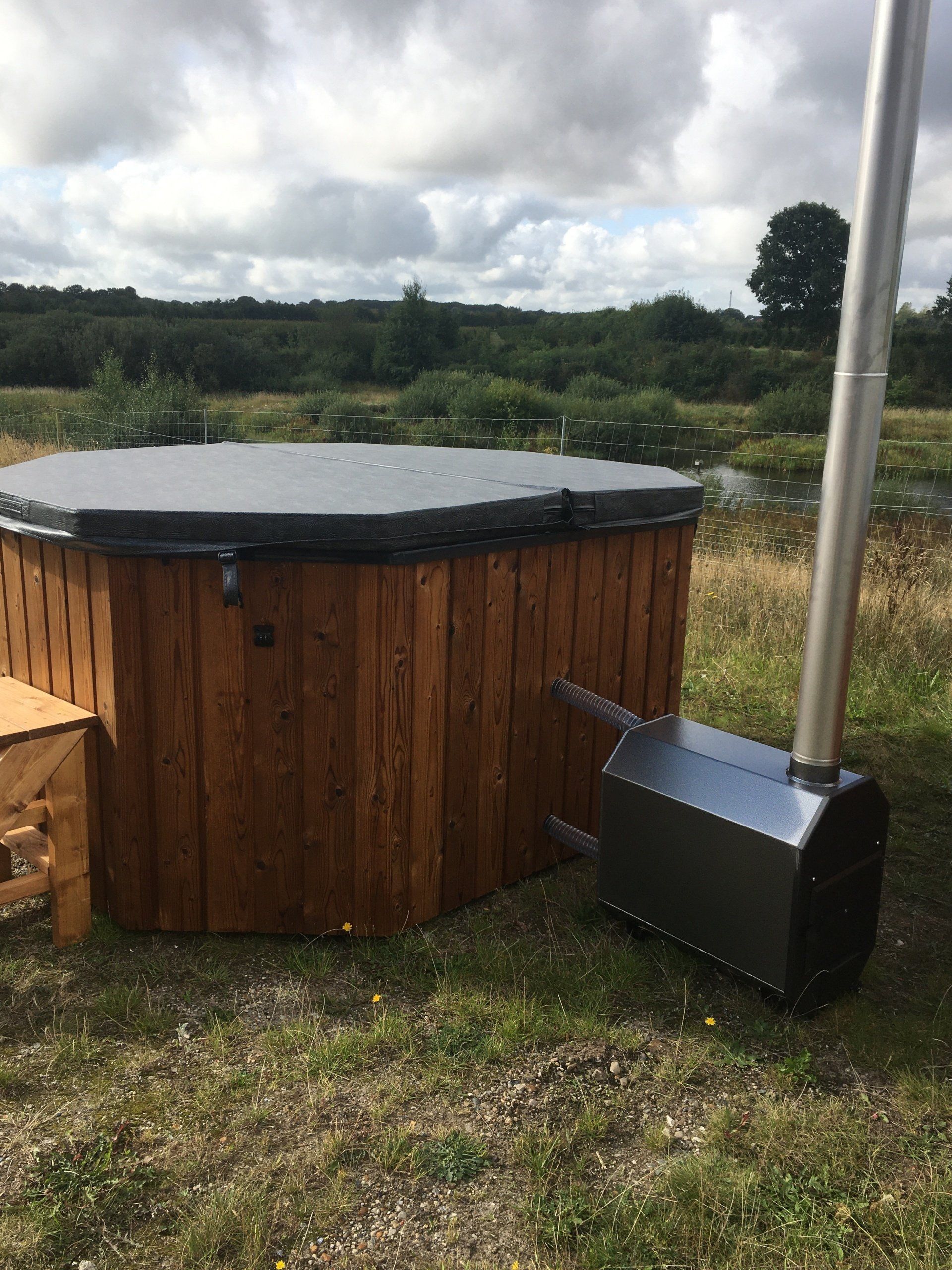 Rundt træjacuzzi med sort låg ved siden af ​​et metalrør til ovnen på en græsklædt udendørs mark.