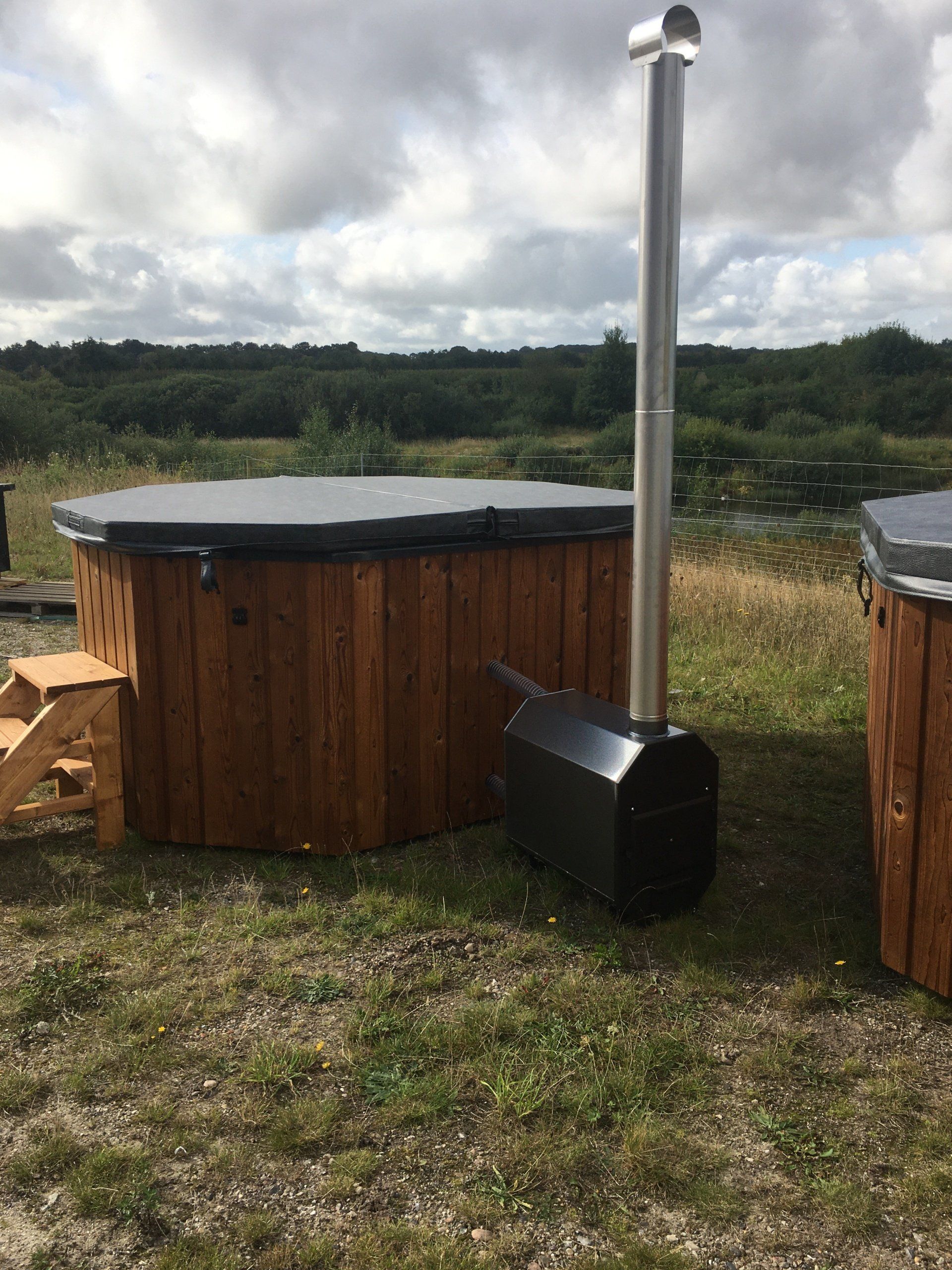 Rundt træjacuzzi med metalskorsten i græsmark under overskyet himmel