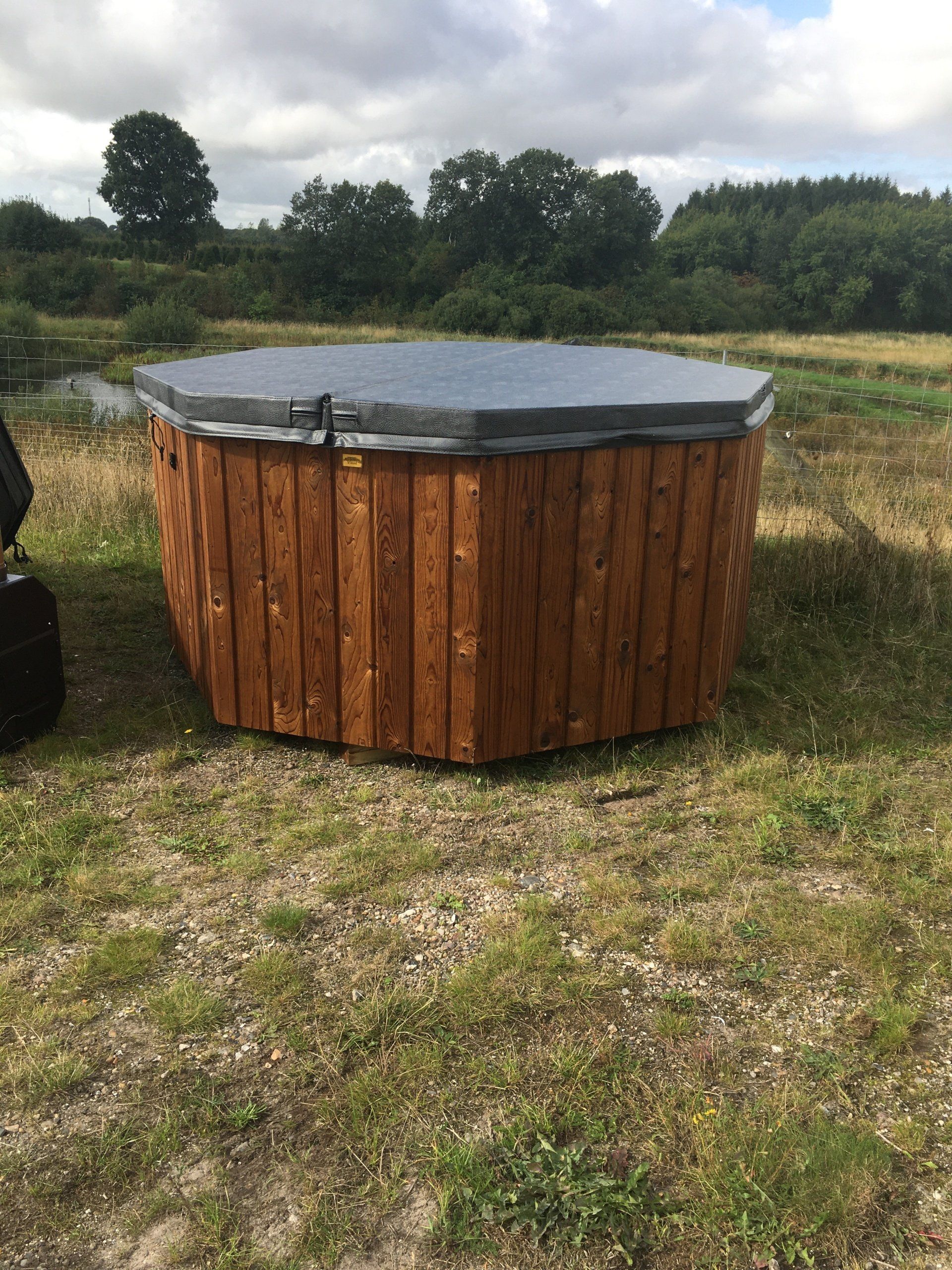 Rundt træjacuzzi med mørkt overtræk i en græsklædt mark under overskyet himmel.