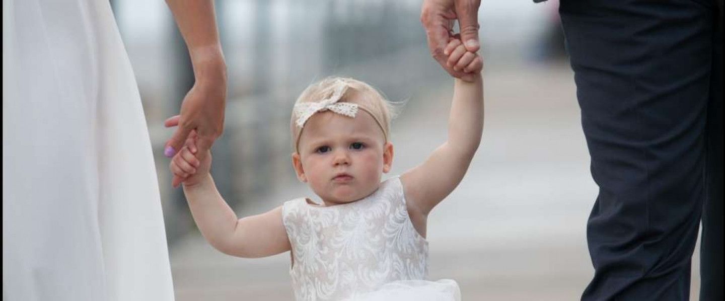 A little girl in a white dress is holding the hands of a bride and groom.