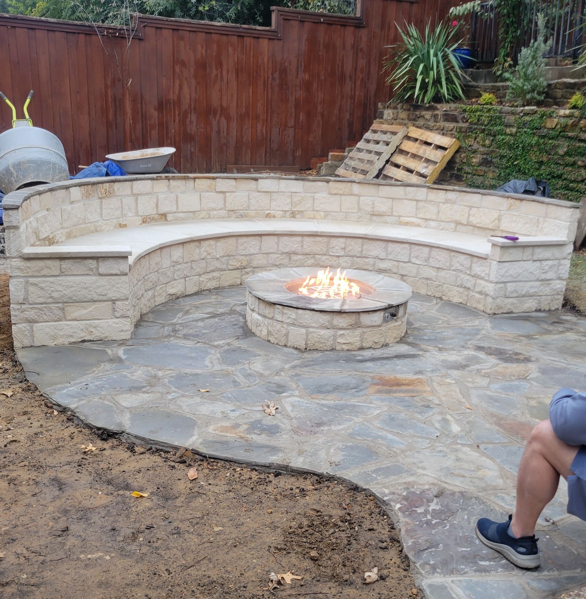 A curved stone seating wall encircling a lit fire pit on a stone patio in front of a wooden fence.