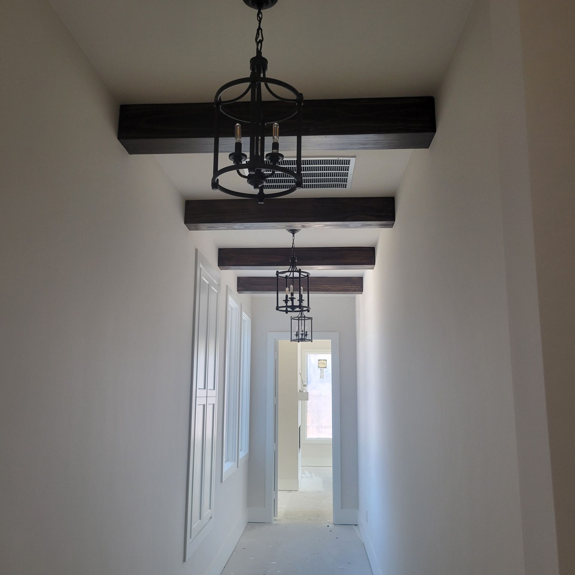 A hallway with white walls and ceiling features dark wooden beams and two hanging black lantern-style pendant lights.