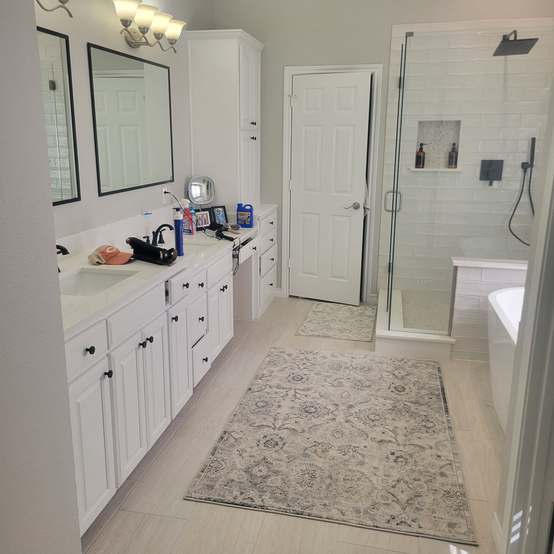 A modern white bathroom with double vanity, mirrors, a glass-enclosed shower, a white door, and a patterned area rug.