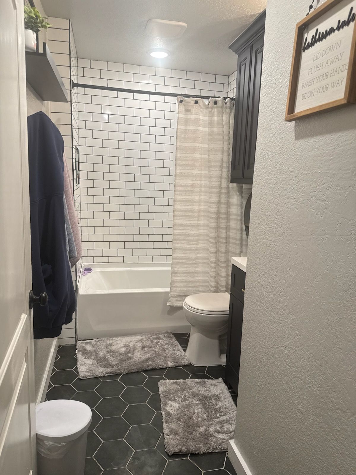 A small bathroom featuring black hexagonal floor tiles, a white brick-patterned tub surround, and a dark wood vanity.