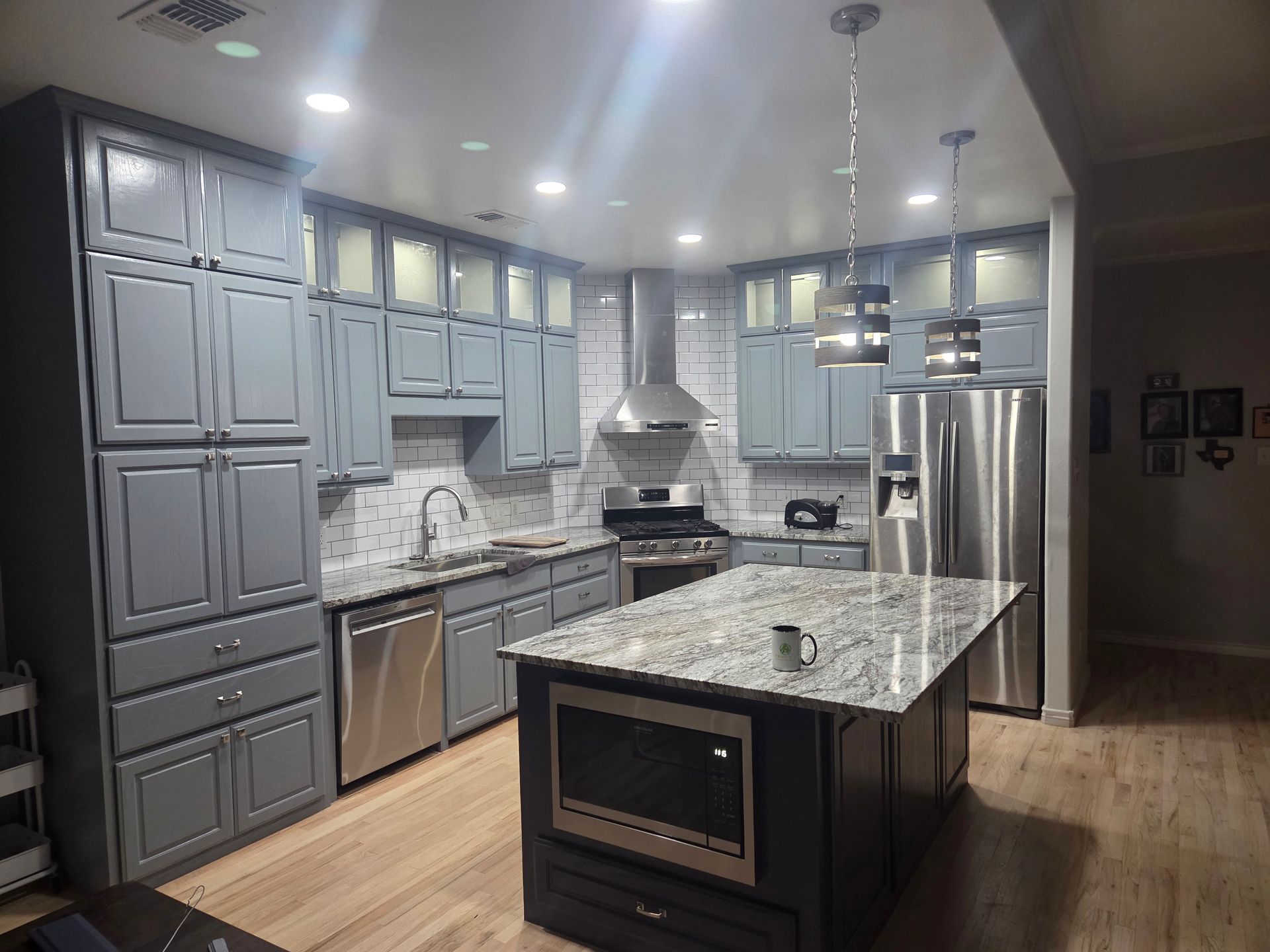 A modern kitchen with grey cabinets, granite countertops, a central island with a built-in microwave, and light wood floors.