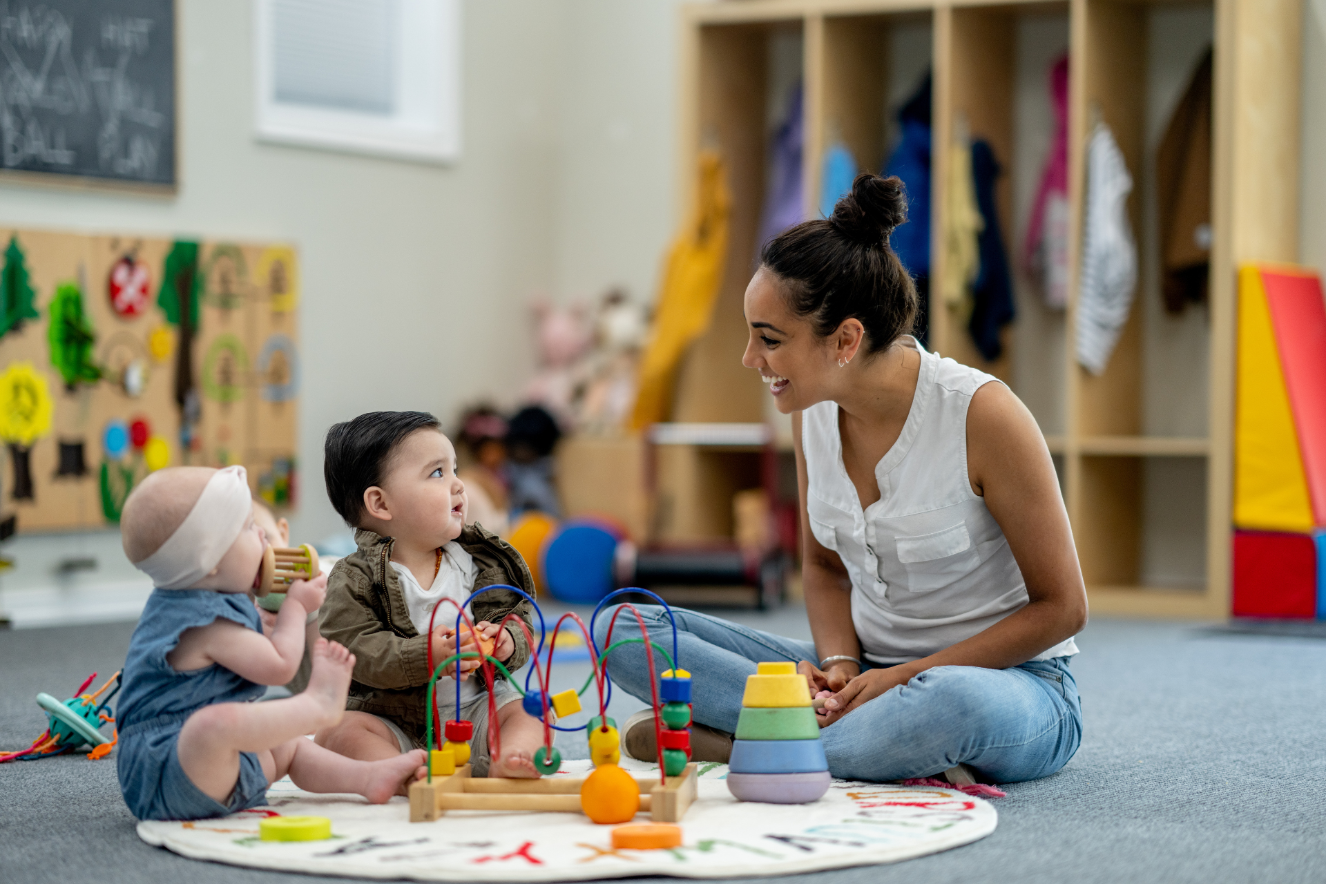 Teacher and two toddlers play with colorful toys on a classroom rug.