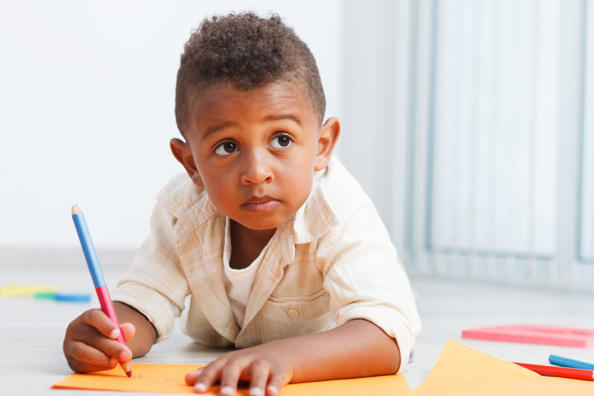 Child drawing with colored pencils at a table, looking up in a bright classroom.