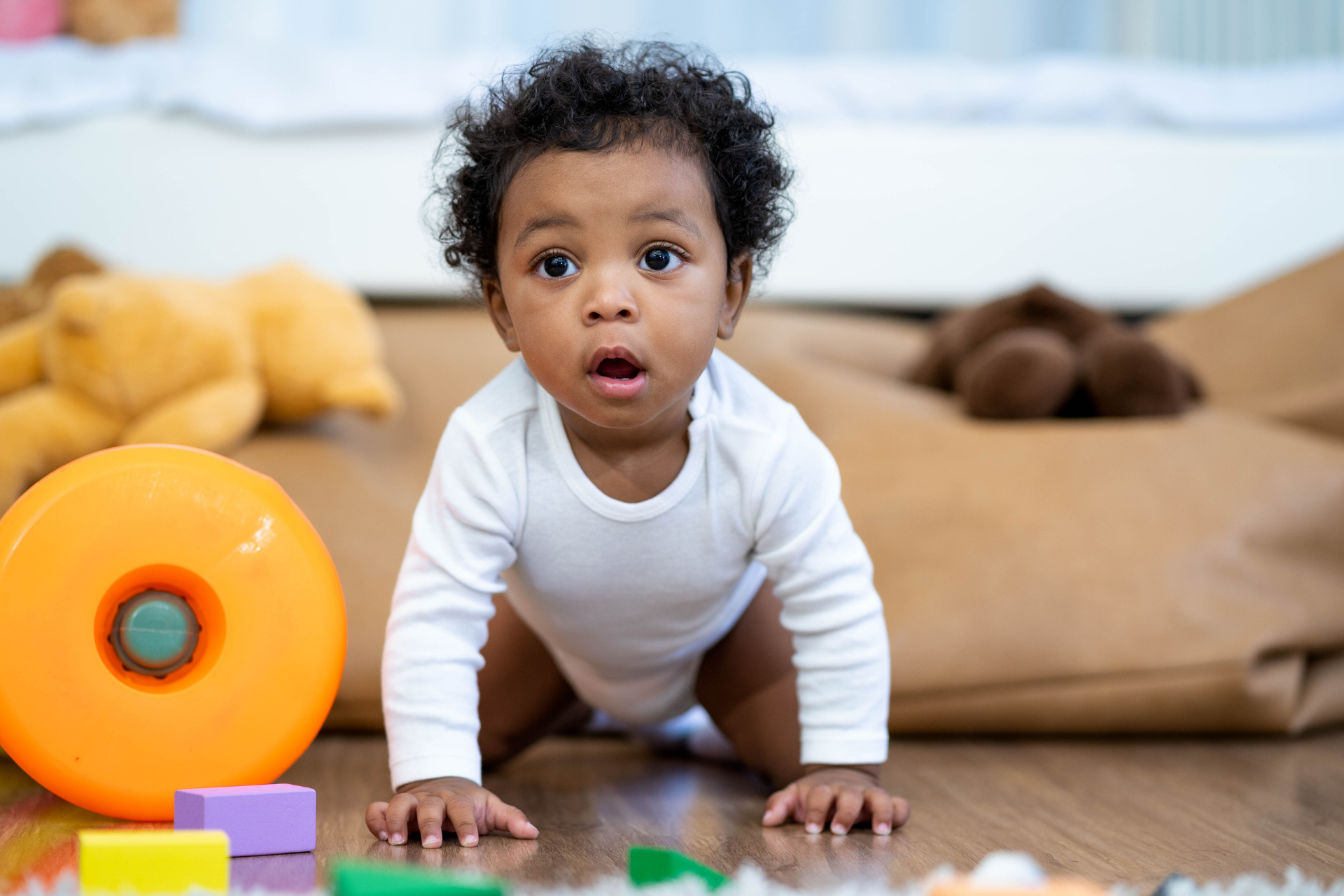 Baby crawling on floor beside orange ring toy in a playroom, with stuffed animals in the background