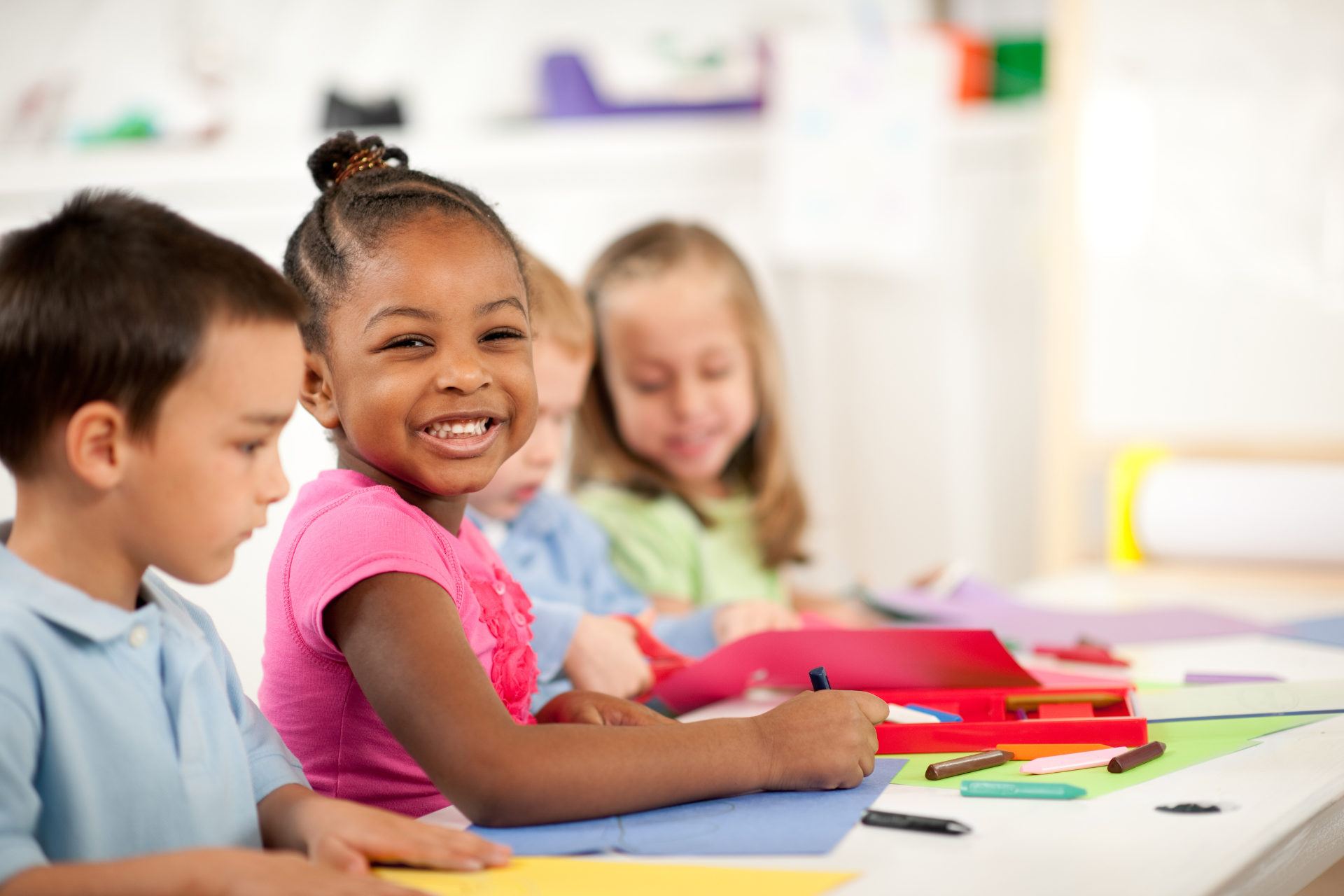 Children sitting at a classroom table, smiling and drawing with colorful art supplies
