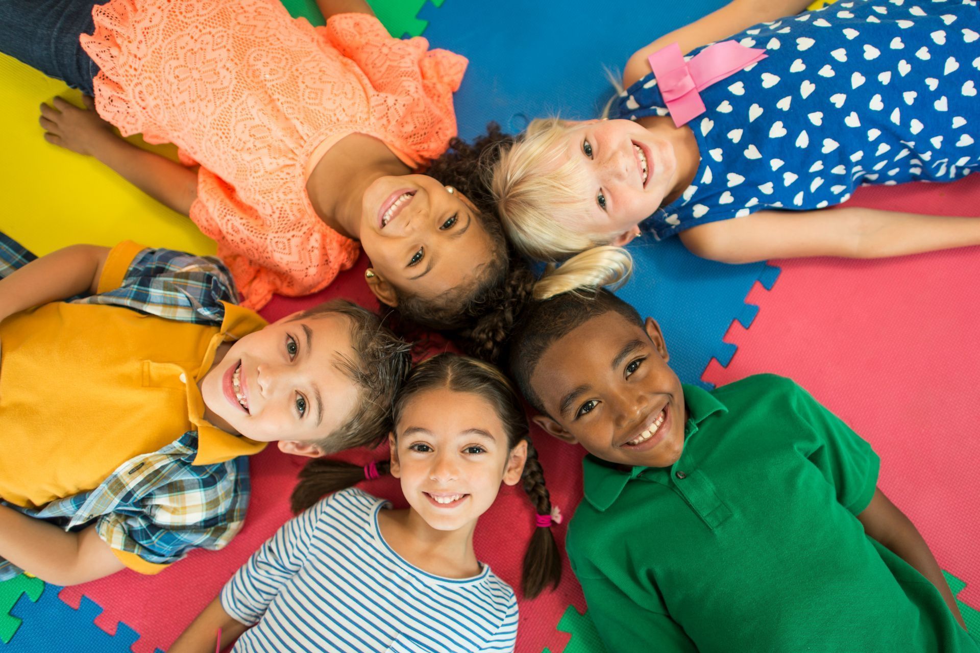 Five children lying in a circle on colorful puzzle mats, smiling at the camera