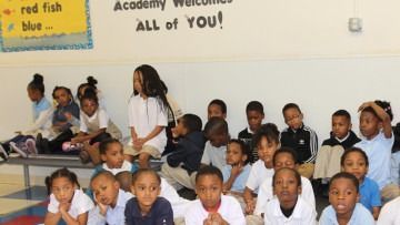 Group of children seated in a classroom, with a teacher standing behind them and a “Welcome” sign on the wall.