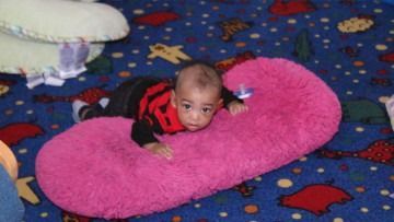 Baby lying on a bright pink plush cushion on a patterned blue carpet
