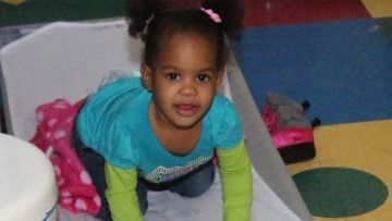 Toddler kneeling on a colorful classroom mat and smiling at the camera