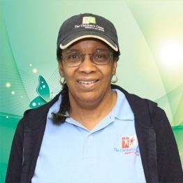 Woman in a black cap and glasses, smiling in a light blue polo against a green gradient background.