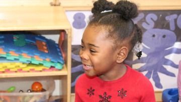 Smiling child in a red shirt in a colorful classroom, looking to the side.