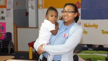 Adult holding a toddler in a classroom, both facing the camera and smiling.