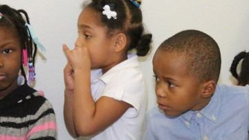 Three children sitting side by side, one holding a hand to their face, against a plain white wall
