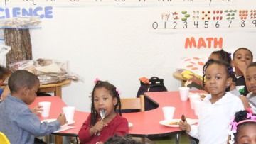 Children sitting at red tables in a classroom, with science and math posters on the wall.